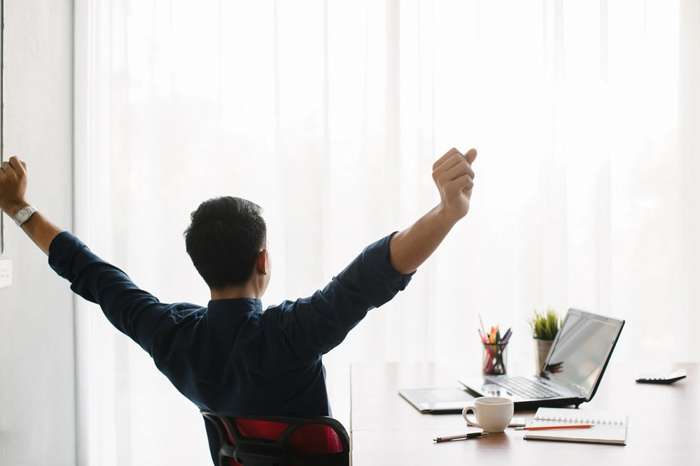 © uinmine / Adobe Stock Ein Mann sitzt am Computer und dehnt und streckt sich.